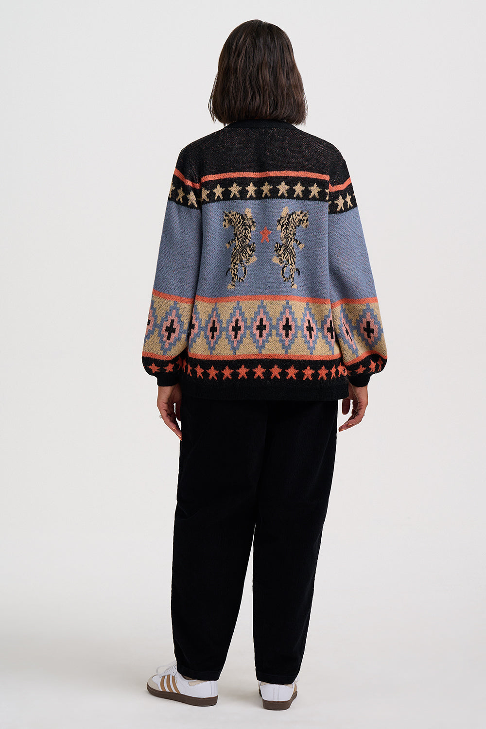 A person shown from the back wearing a multicolor knit sweater with geometric patterns and embroidered horse motifs, paired with black pants and white sneakers, against a plain light gray studio background.