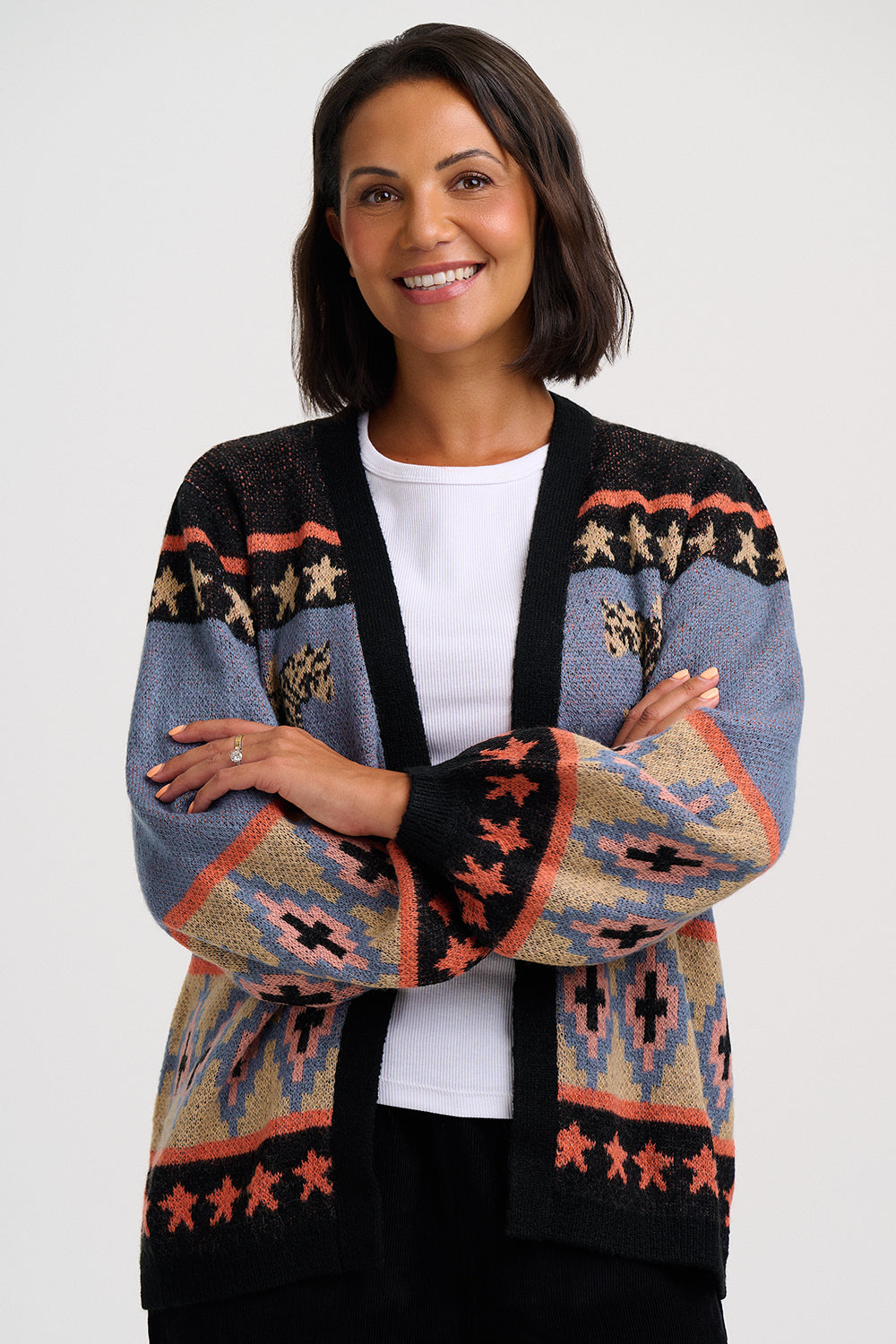 A smiling woman with short dark hair models a knit cardigan with a Southwestern pattern. The cardigan features a mix of slate blue, black, beige, and terracotta colors in geometric and star motifs, worn over a white t-shirt and black pants against a light gray studio background.
