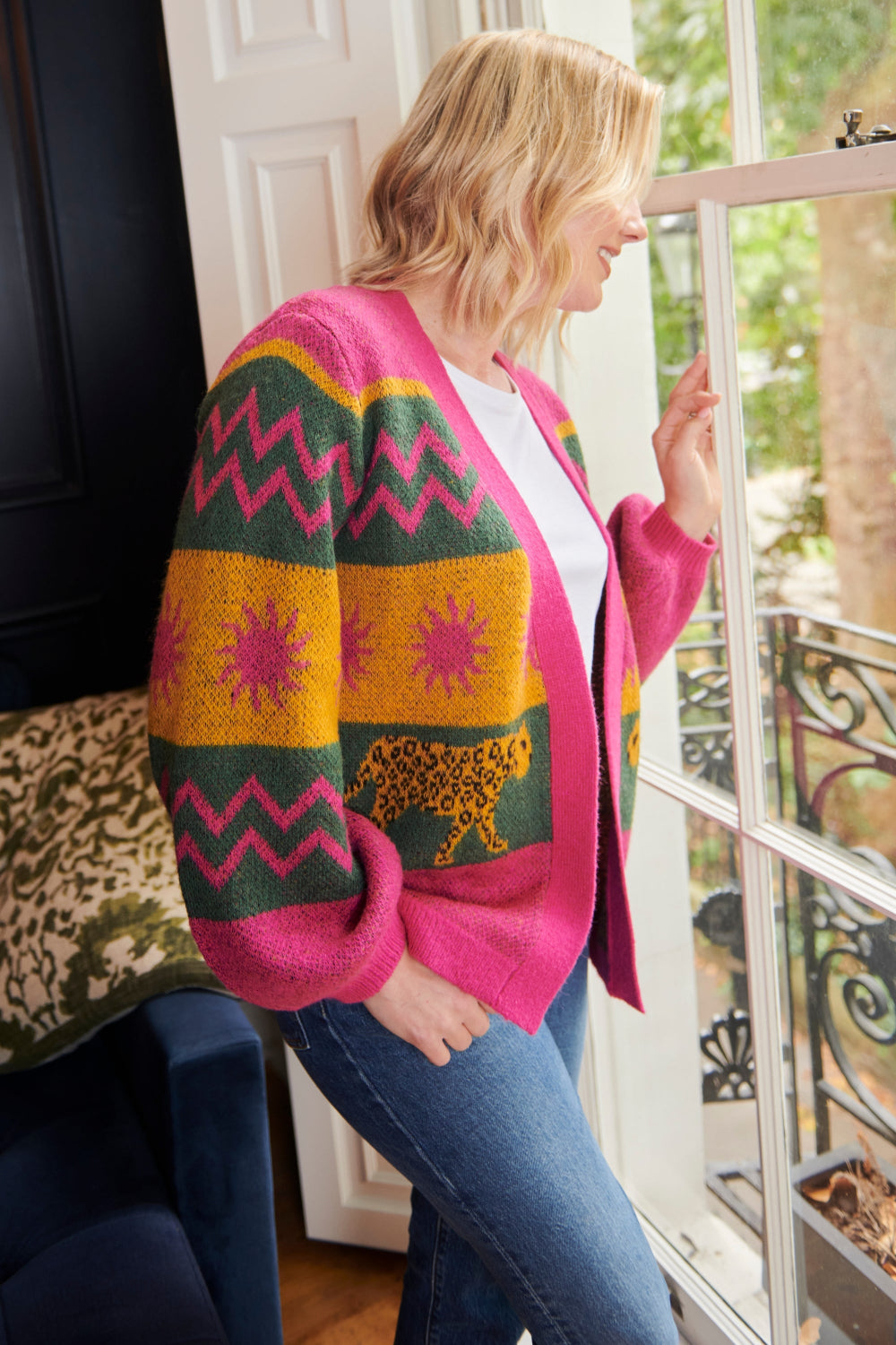 A woman standing by a window wearing a multicolored cardigan with sun leopards pattern, balloon sleeves, and a relaxed fit.