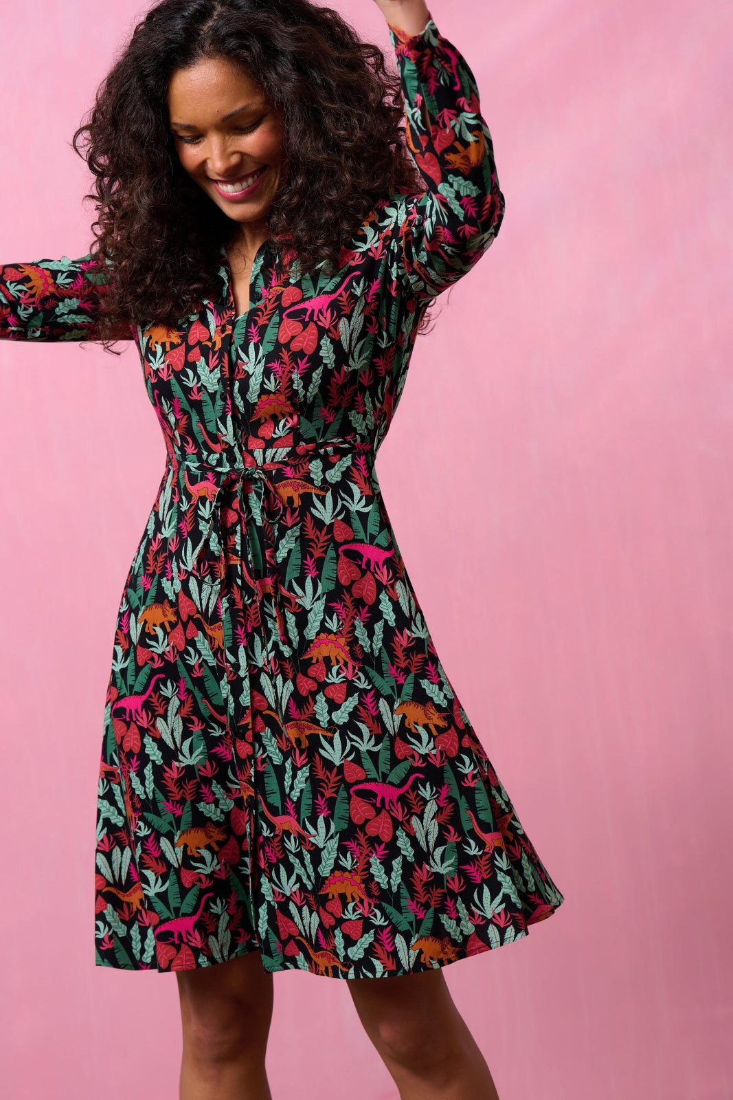 A woman is posing and smiling, wearing a black shirt dress with a dinosaur print.