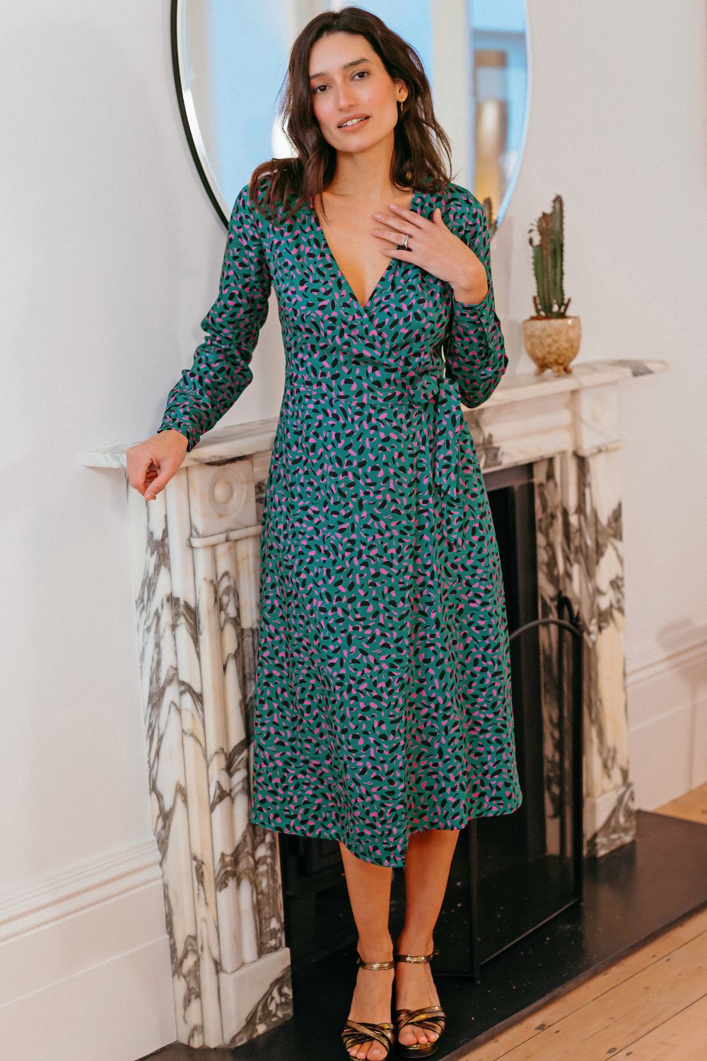 Woman in a green floral dress standing in a room with a fireplace and cactus.