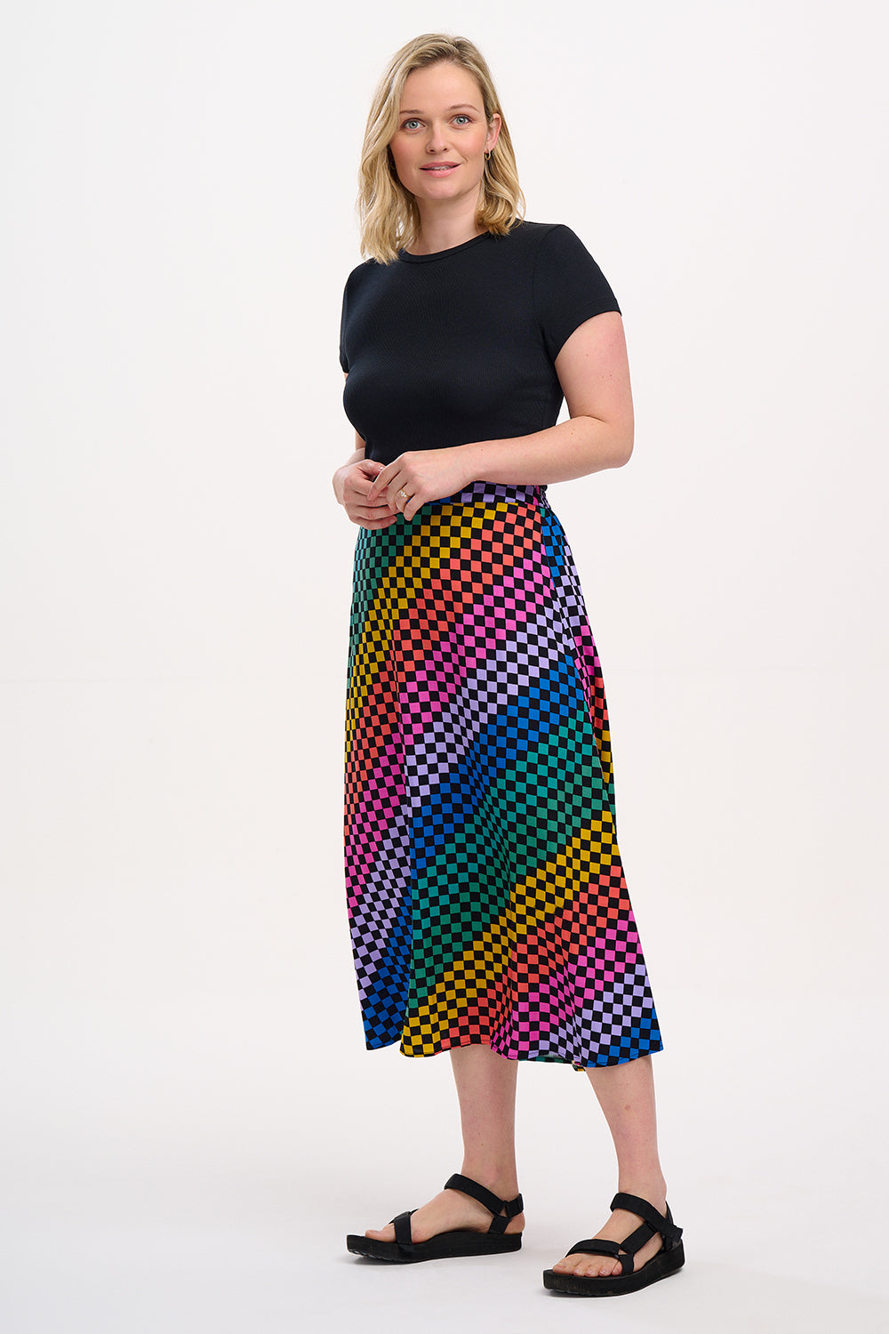 A woman standing and wearing a high-waisted, midi-length Zora skirt with a Rainbow Checkerboard design on a black base.