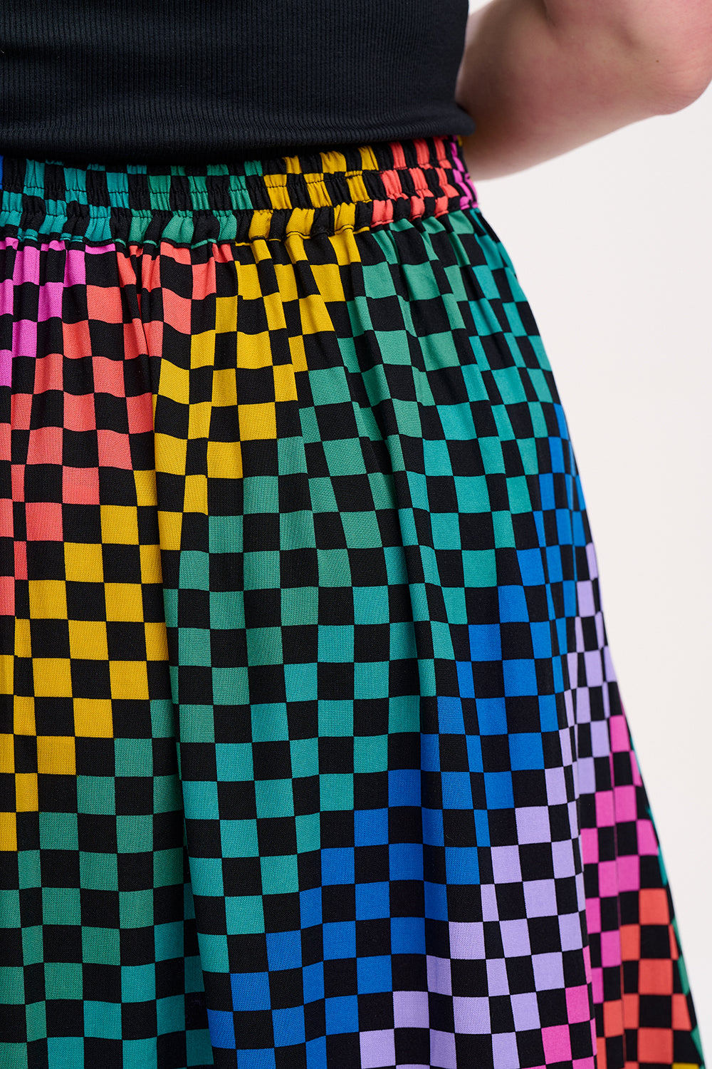 A woman standing and wearing a high-waisted, midi-length Zora skirt with a Rainbow Checkerboard design on a black base.