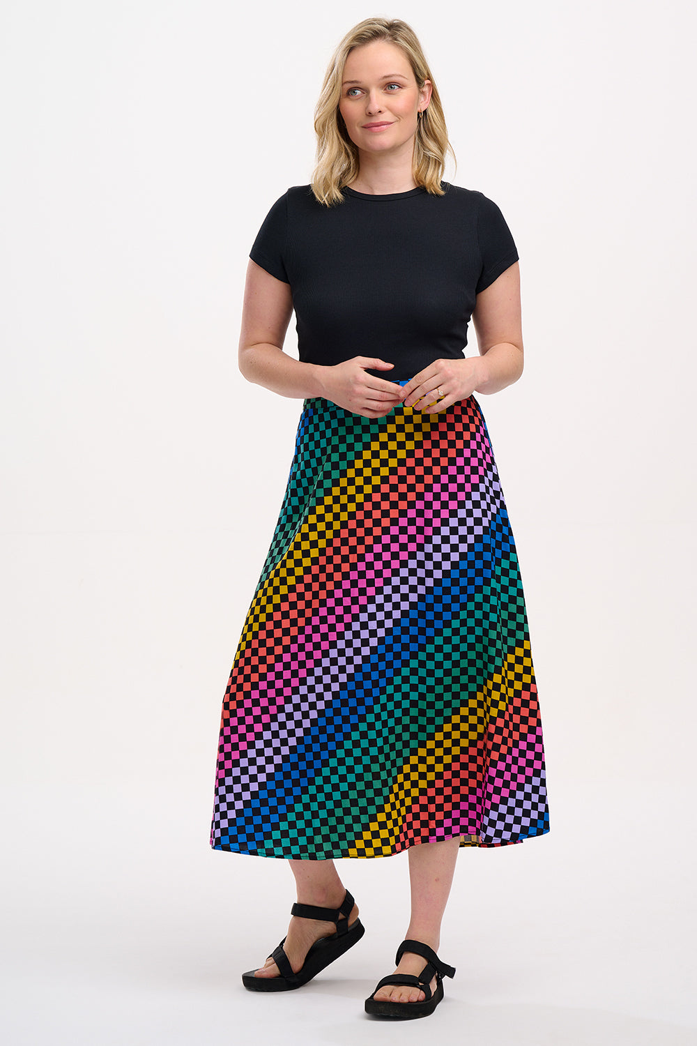 A woman standing and wearing a high-waisted, midi-length Zora skirt with a Rainbow Checkerboard design on a black base.