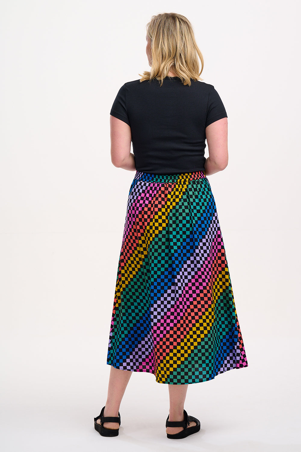 A woman standing and wearing a high-waisted, midi-length Zora skirt with a Rainbow Checkerboard design on a black base.