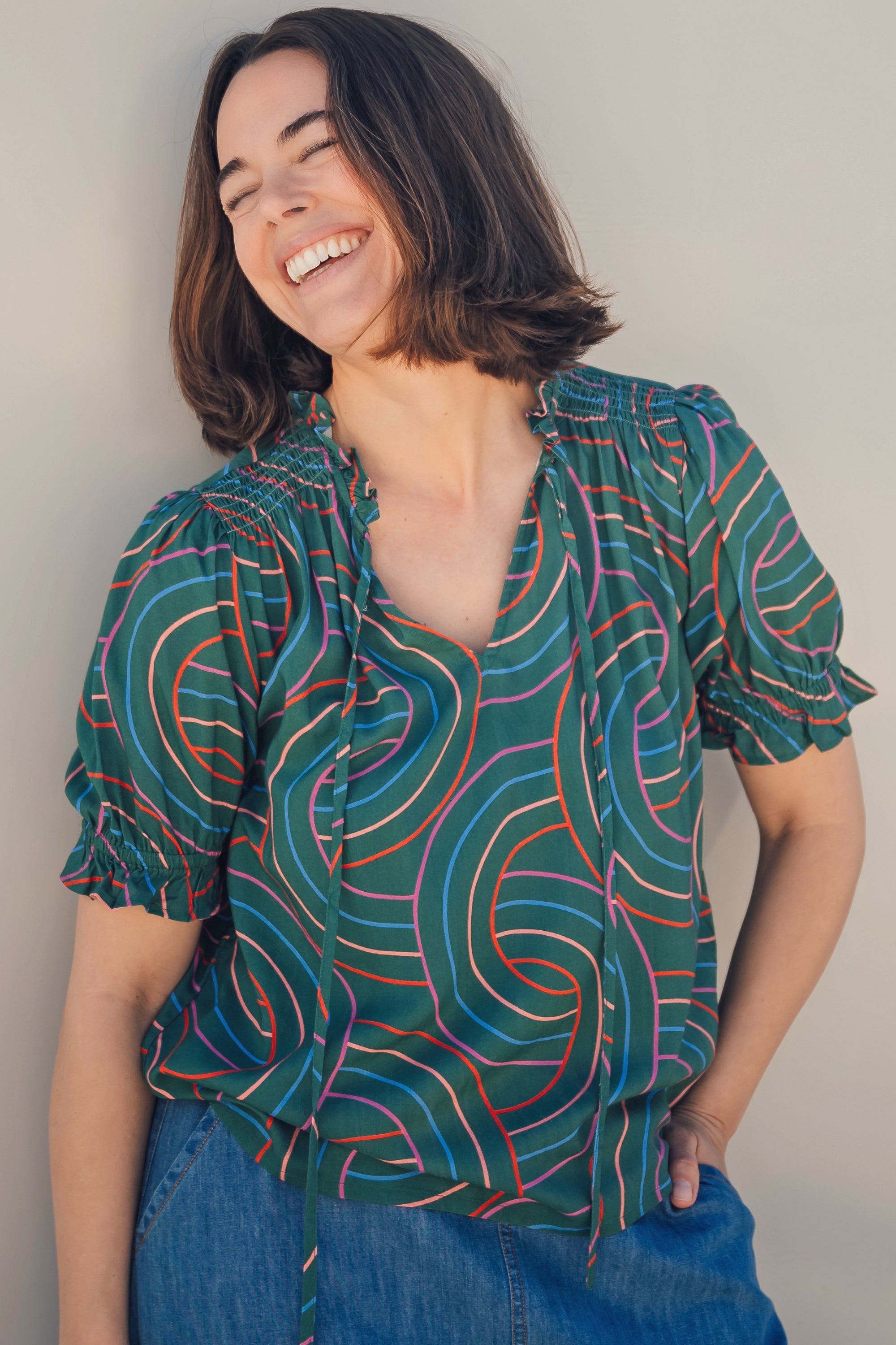 A woman smiling and wearing a green blouse with a geometric concentric circles pattern, featuring a frill collar and v-neckline with ties, short puff sleeves with elasticated frill cuffs.