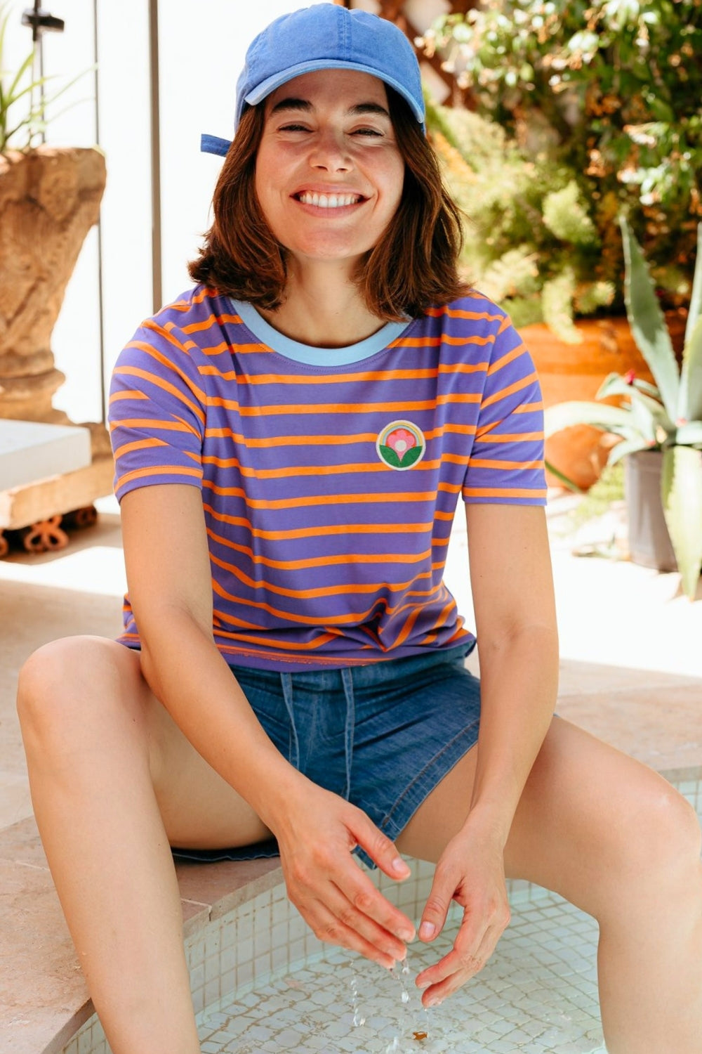 Woman wearing a striped shirt with a logo and denim shorts, sitting outdoors.