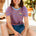 Woman wearing a striped shirt with a logo and denim shorts, sitting outdoors.