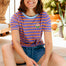 Woman wearing a striped shirt with a logo and denim shorts, sitting outdoors.
