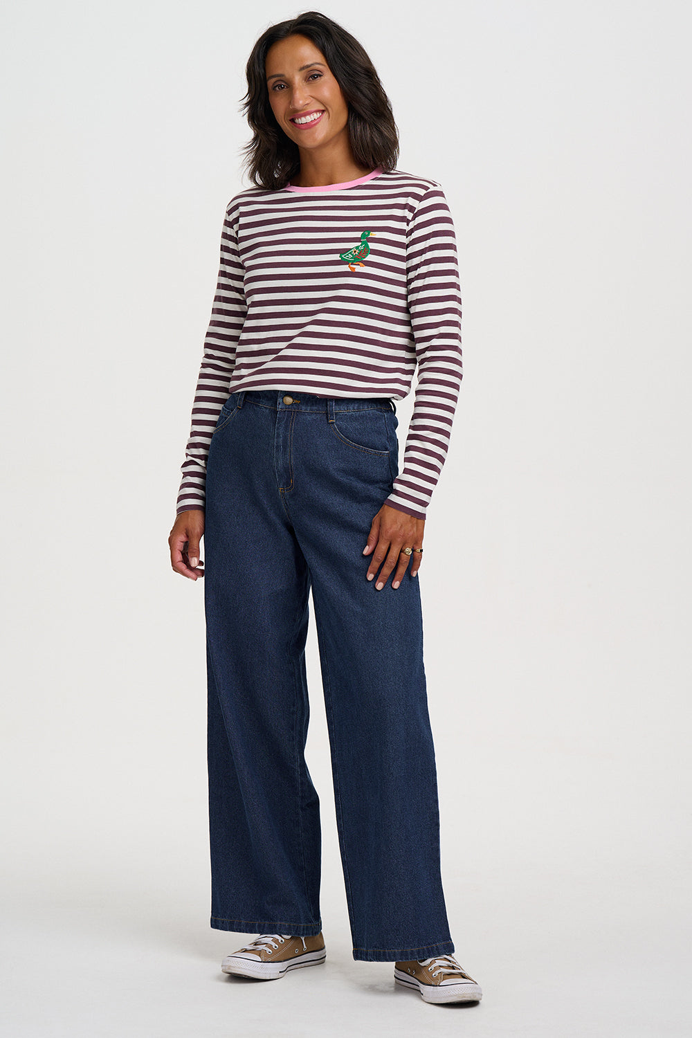 A woman modeling a long-sleeve striped shirt with a small green embroidered bird on the chest, paired with dark blue wide-leg jeans and brown sneakers, against a plain light gray studio background.