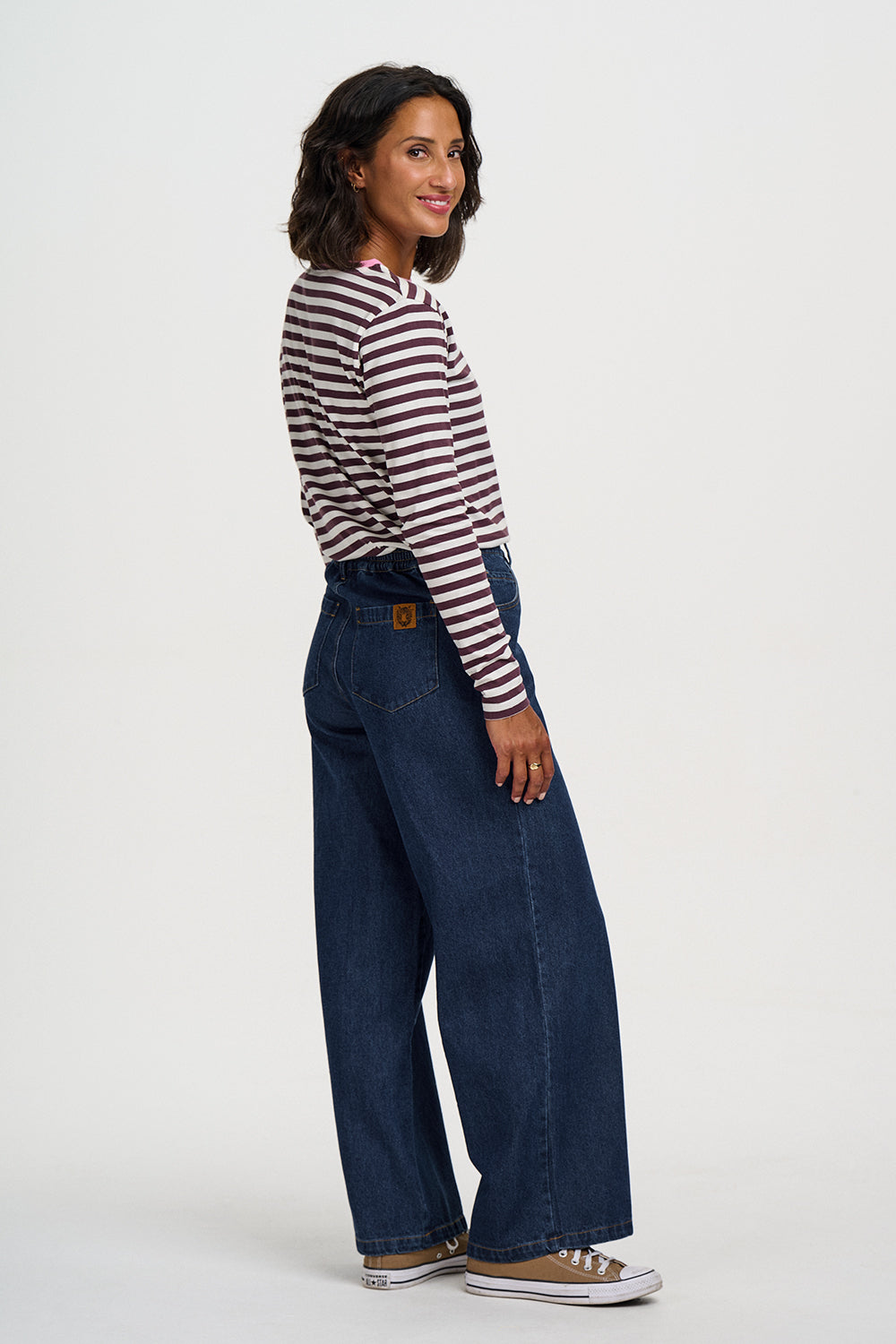 A woman modeling a pair of dark blue wide-leg denim jeans with a striped long-sleeve shirt, standing against a plain light gray studio background.