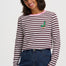 A woman wearing a long-sleeve crop top with dark red and white horizontal stripes, a light pink crew neck, and a small embroidered green duck on the chest, paired with dark blue jeans against a light gray background.