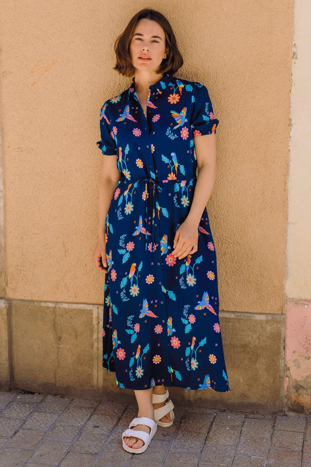 Woman wearing a blue floral dress standing against a beige wall.