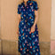 Woman wearing a blue floral dress standing against a beige wall.