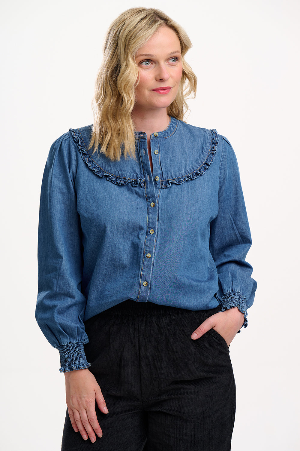 Sugarhill Brighton model wearing a blue denim blouse with ruffled details on a white background