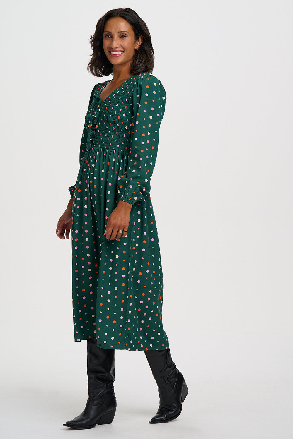 A woman modeling a dark green midi dress with a colorful polka dot pattern, paired with black knee-high boots, against a plain light gray studio background.