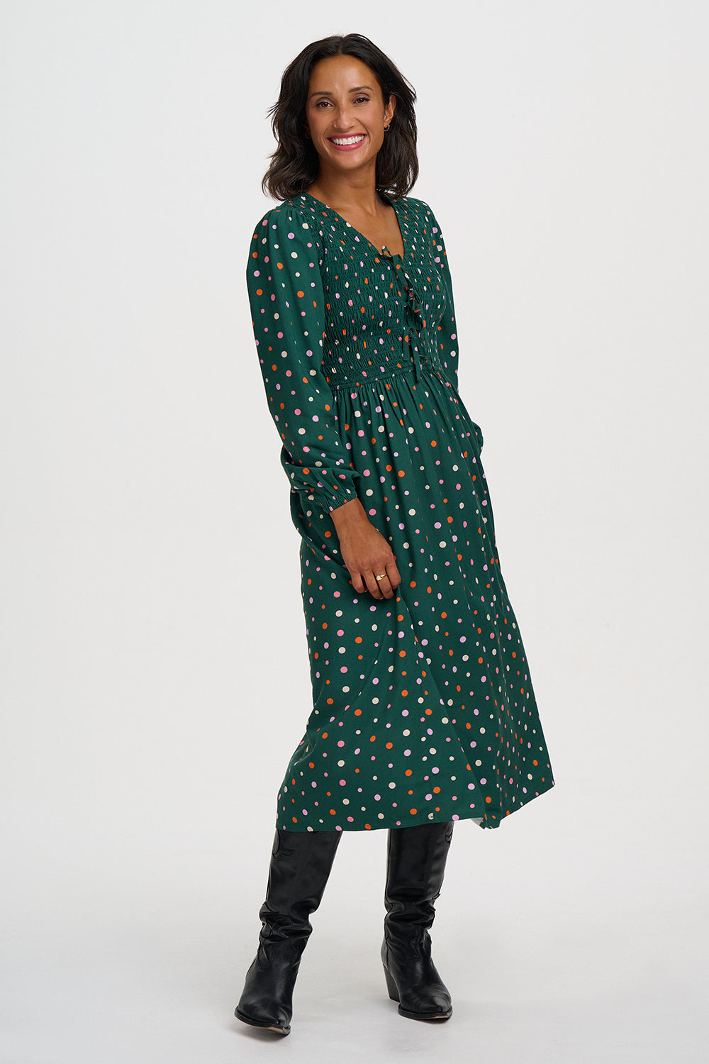 A woman modeling a dark green long-sleeve midi dress with a white and orange polka dot pattern, paired with black knee-high boots, against a plain light gray studio background.