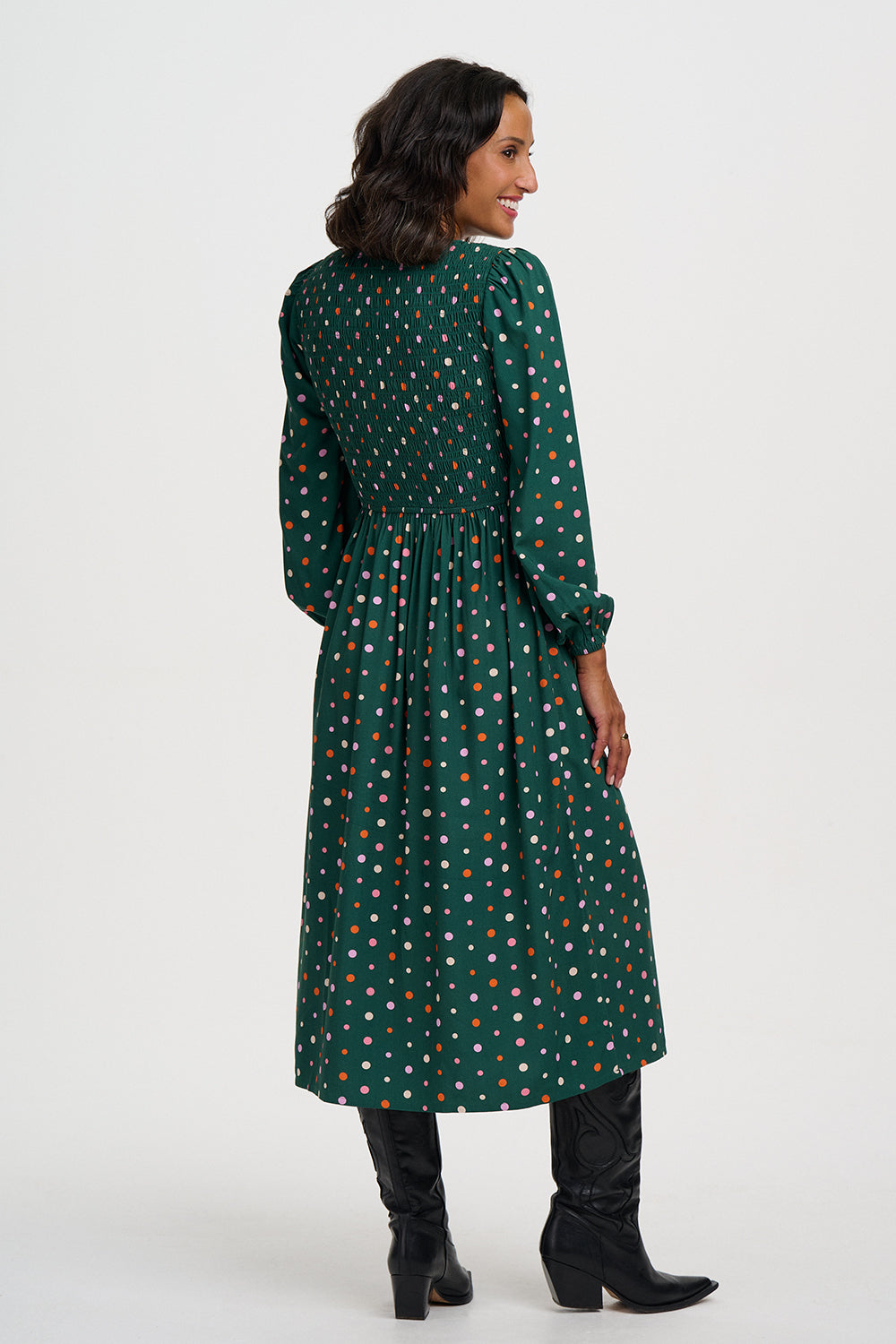 A woman shown from the back wearing a dark green midi dress with a small multicolor polka dot pattern, paired with black knee-high boots, against a plain light gray studio background.