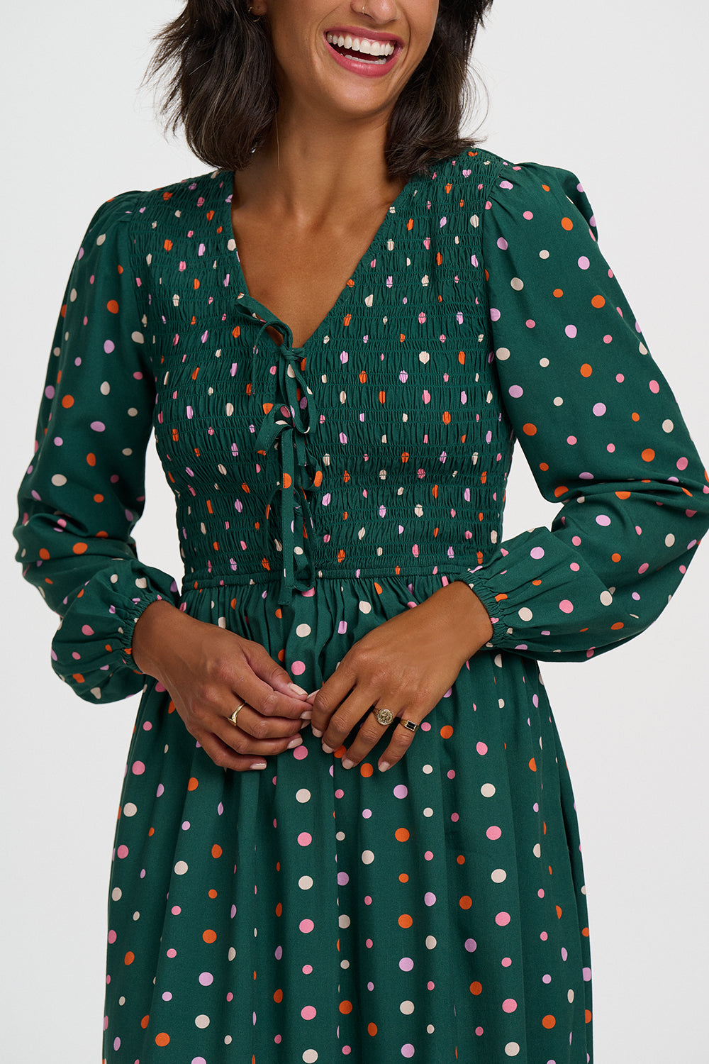 A woman wearing a dark green long-sleeve dress with a colorful polka dot pattern, featuring a smocked bodice and tie-front neckline.