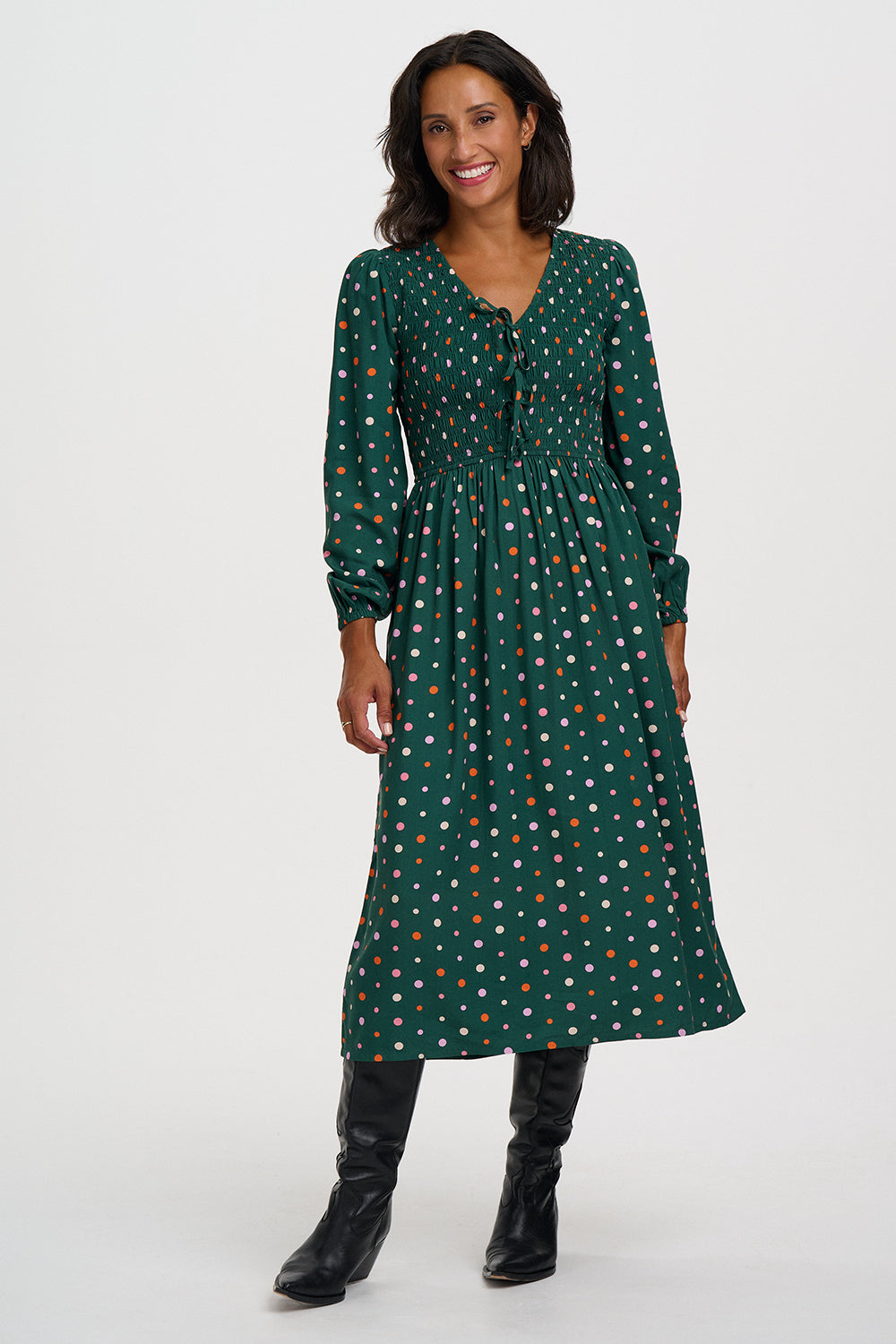 A woman modeling a dark green long-sleeve midi dress with a white and orange polka dot pattern, paired with black knee-high boots, against a plain light gray studio background.