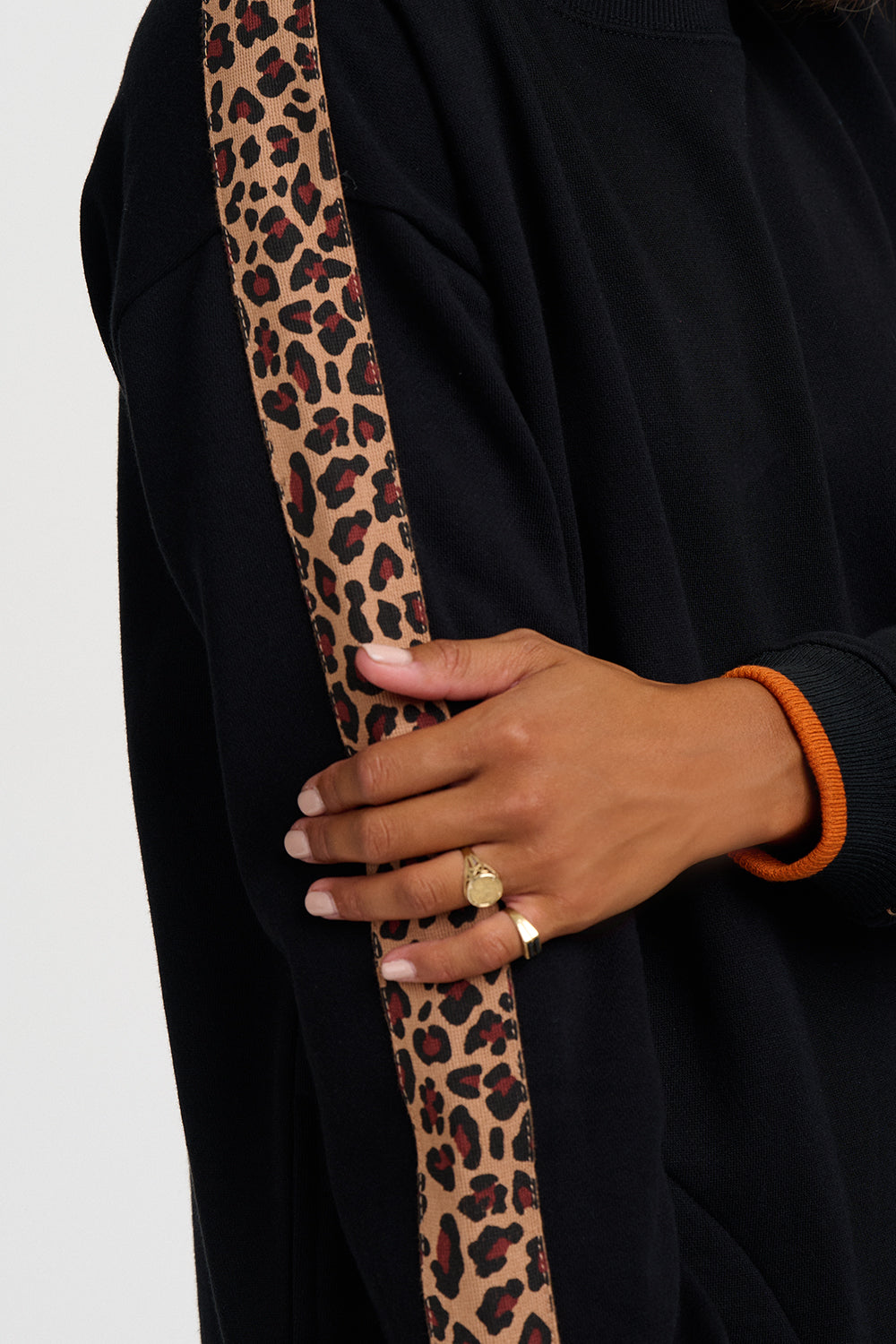Close-up of a person wearing a black sweatshirt with a wide leopard-print stripe running down the sleeve, paired with an orange ribbed cuff and a gold ring.