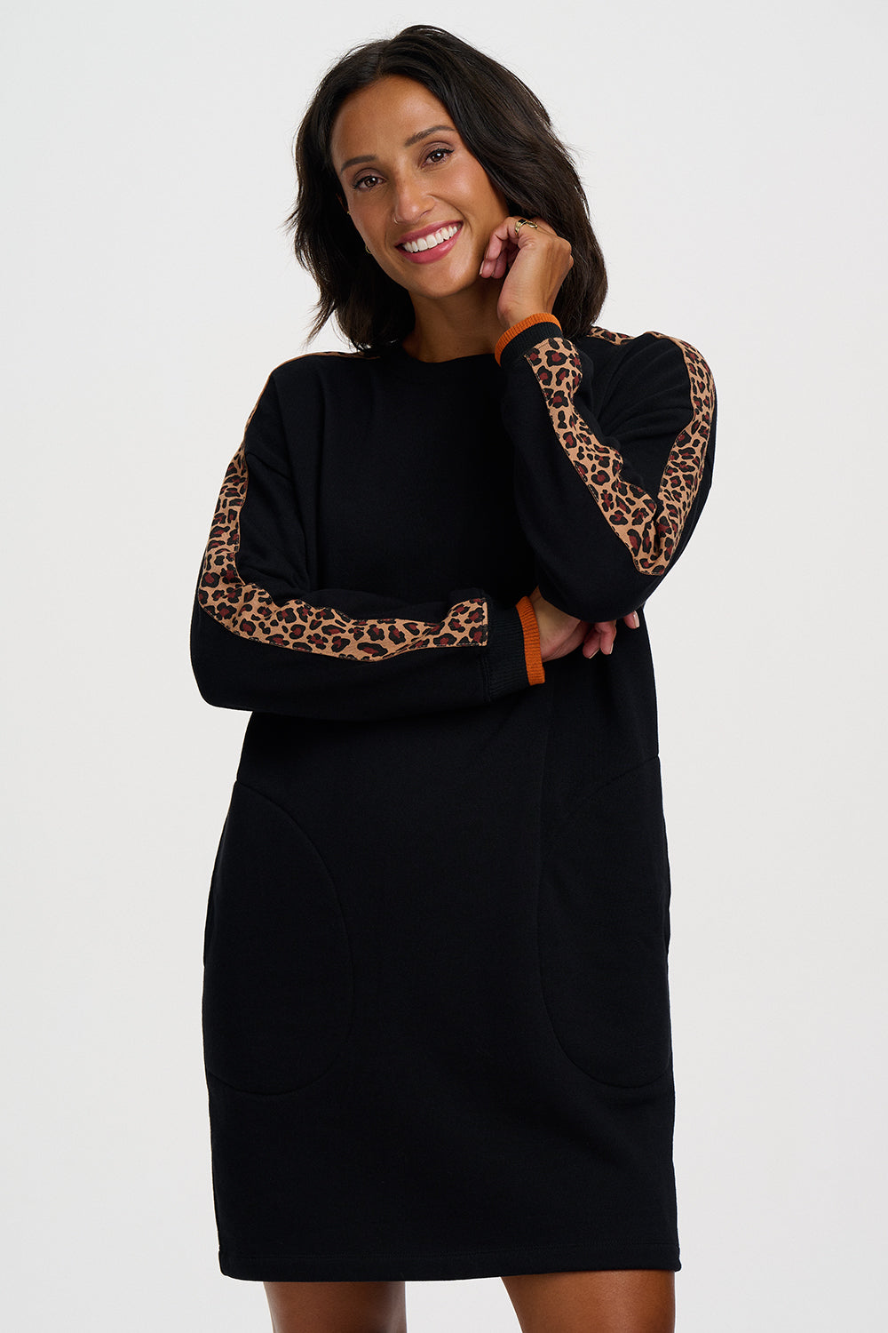 A woman modeling a black long-sleeve dress with leopard print patterns on the sleeves and a small orange-brown accent tab at the cuff, photographed against a plain light gray studio background.