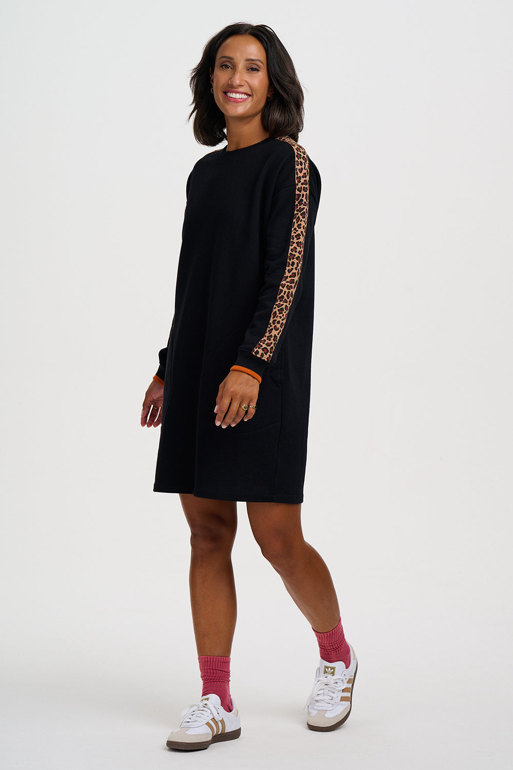 A woman modeling a black long-sleeve mini dress with a leopard-print patterned sleeve, paired with pink socks and white sneakers, against a plain light gray studio background.