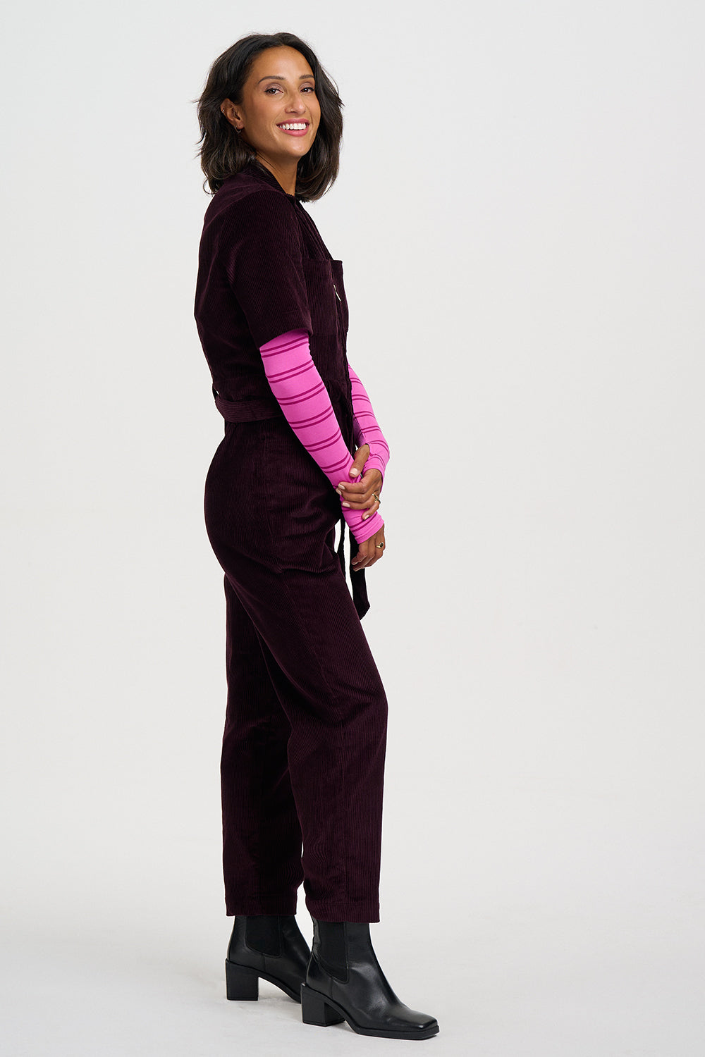 A woman modeling a deep plum velvet jumpsuit with bright pink striped long sleeves, paired with black ankle boots, against a light gray studio background.