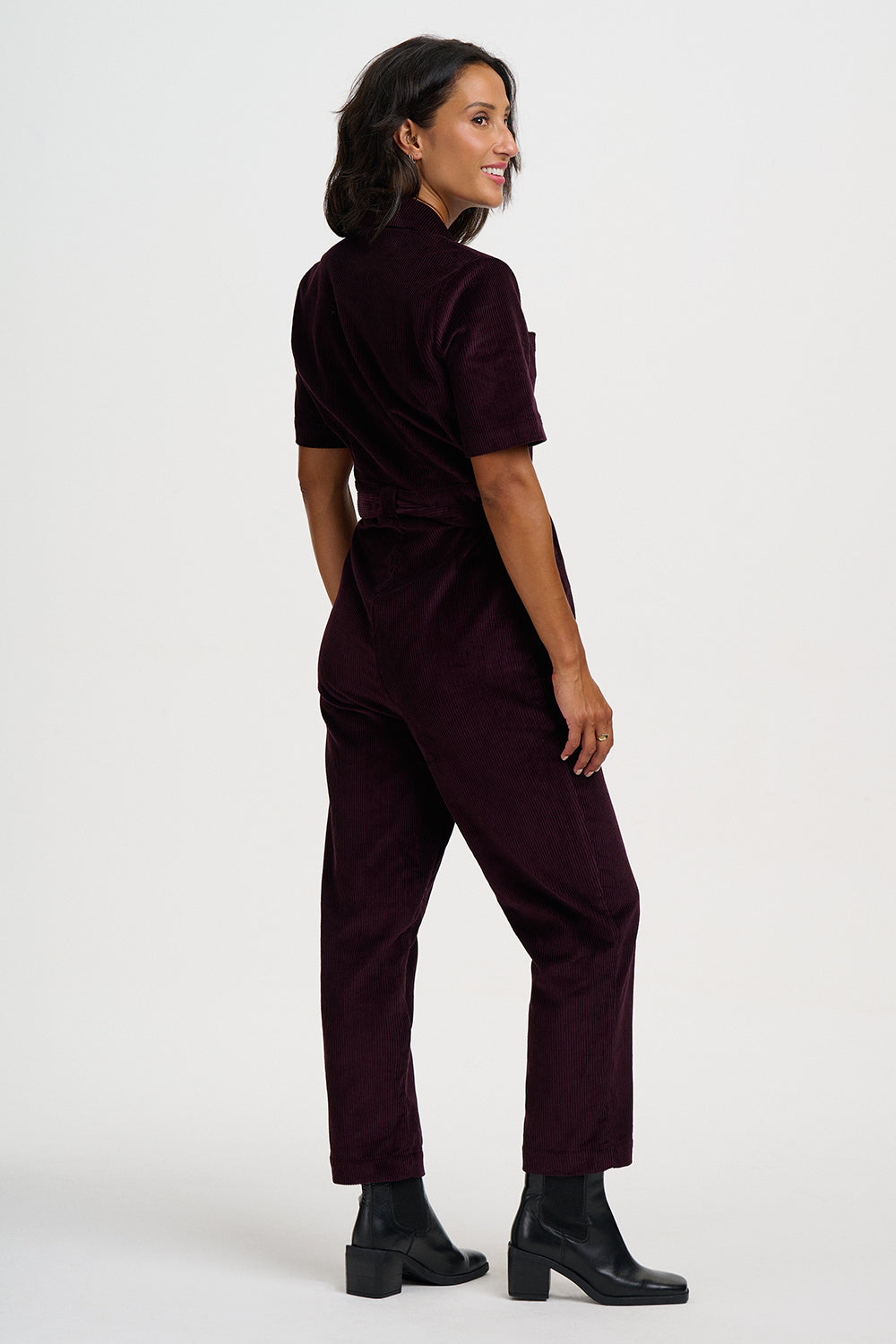 A woman modeling a deep burgundy velvet short-sleeve jumpsuit from the back, paired with black ankle boots, against a plain white studio background.