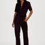A woman modeling a deep plum corduroy jumpsuit with a short zip-up collar, short sleeves, a matching waist tie, and straight legs, paired with black ankle boots against a plain white studio background.