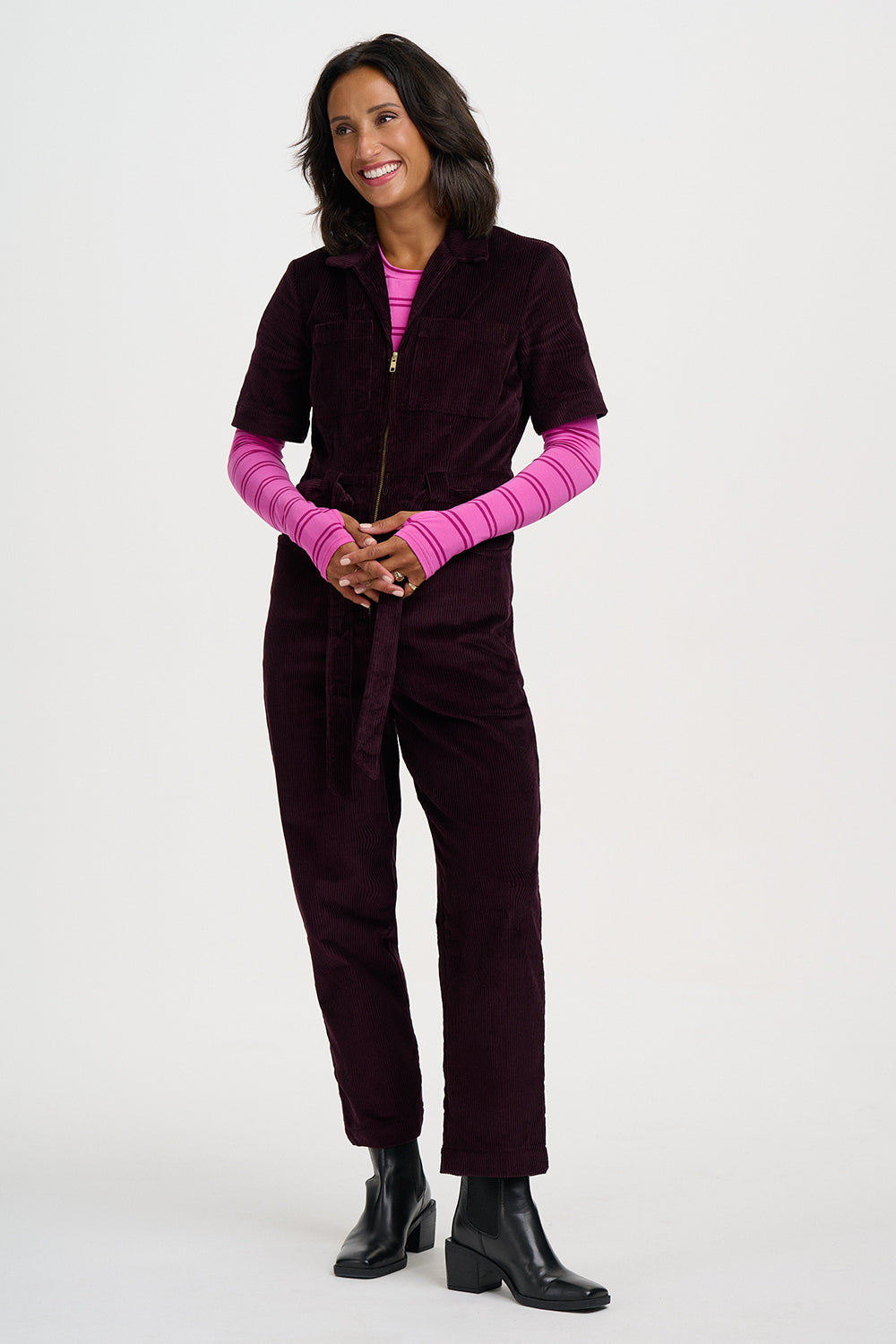 A woman modeling a dark plum corduroy zip-front jumpsuit with short sleeves and a matching waist tie, layered over a bright pink striped long-sleeve top and paired with black ankle boots, on a light gray studio background.