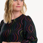 Sugarhill Brighton model wearing a black blouse with colourful patterns on a white background