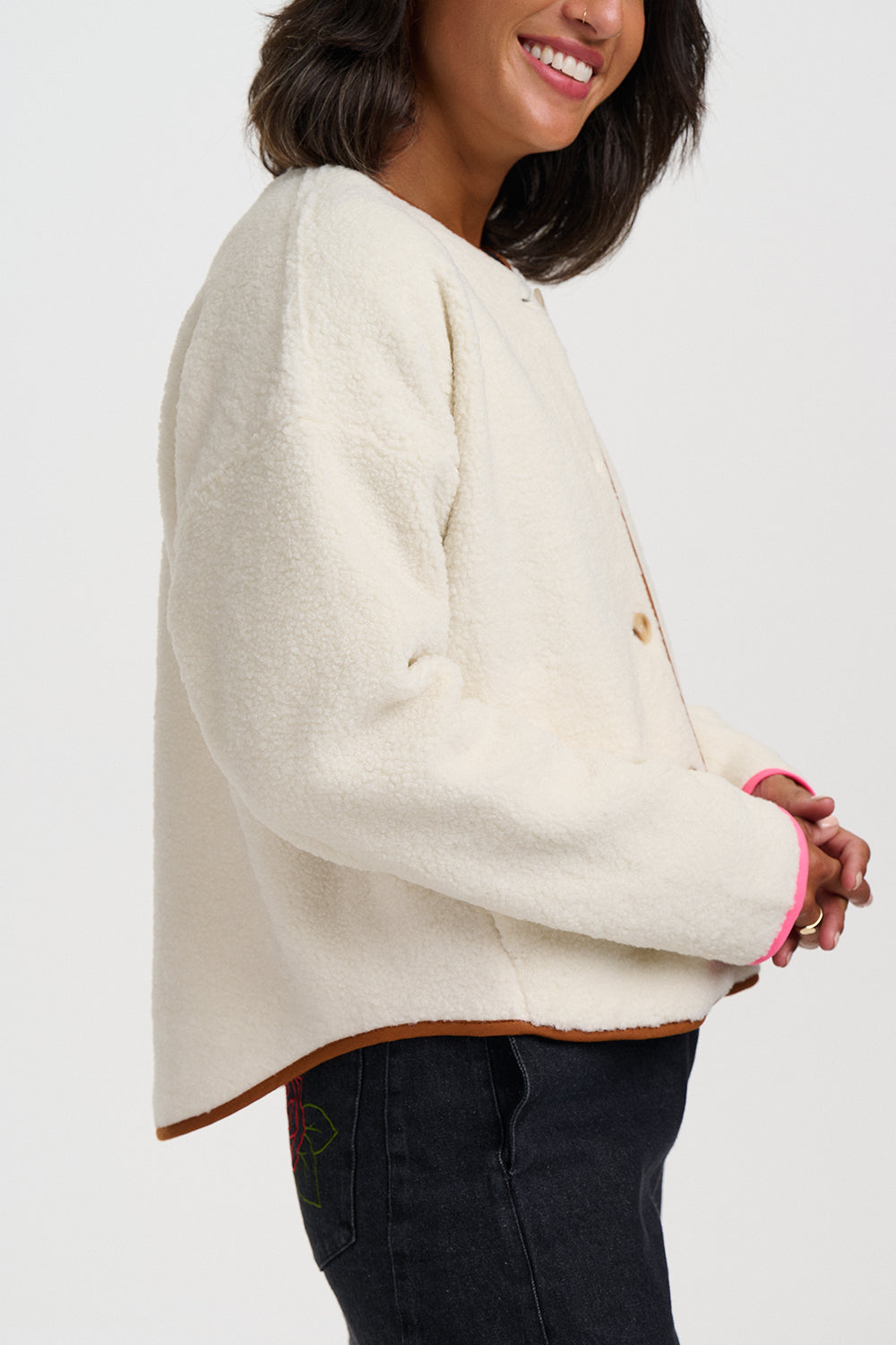 A woman wearing a cream-colored fuzzy cropped jacket with brown trim and a pink cuff accent, paired with dark jeans, shown against a plain light gray background.