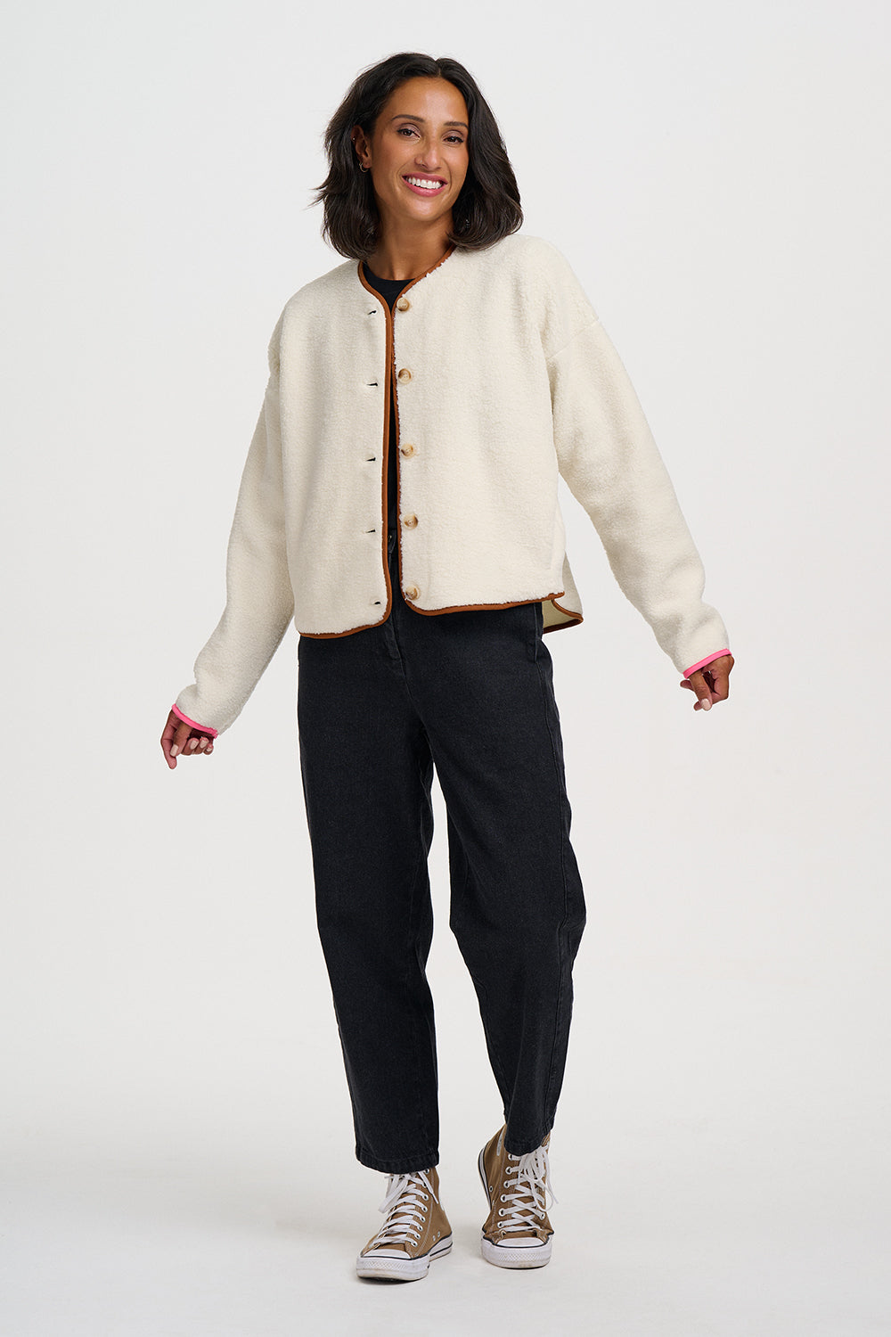 A woman modeling a cream-colored cropped jacket with brown trim and buttons, paired with black pants and beige sneakers, against a plain light gray background.
