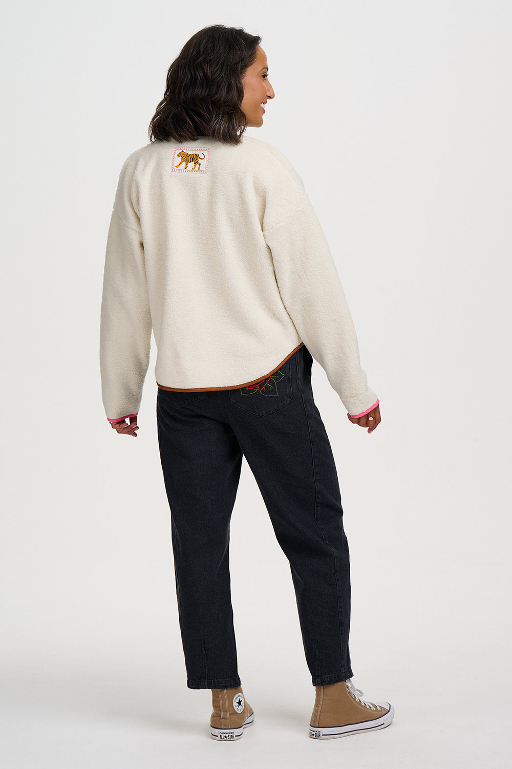 A woman is shown from the back wearing a cream-colored knit sweater with a small embroidered animal patch on the upper back, paired with dark jeans and tan sneakers, against a plain white studio background.
