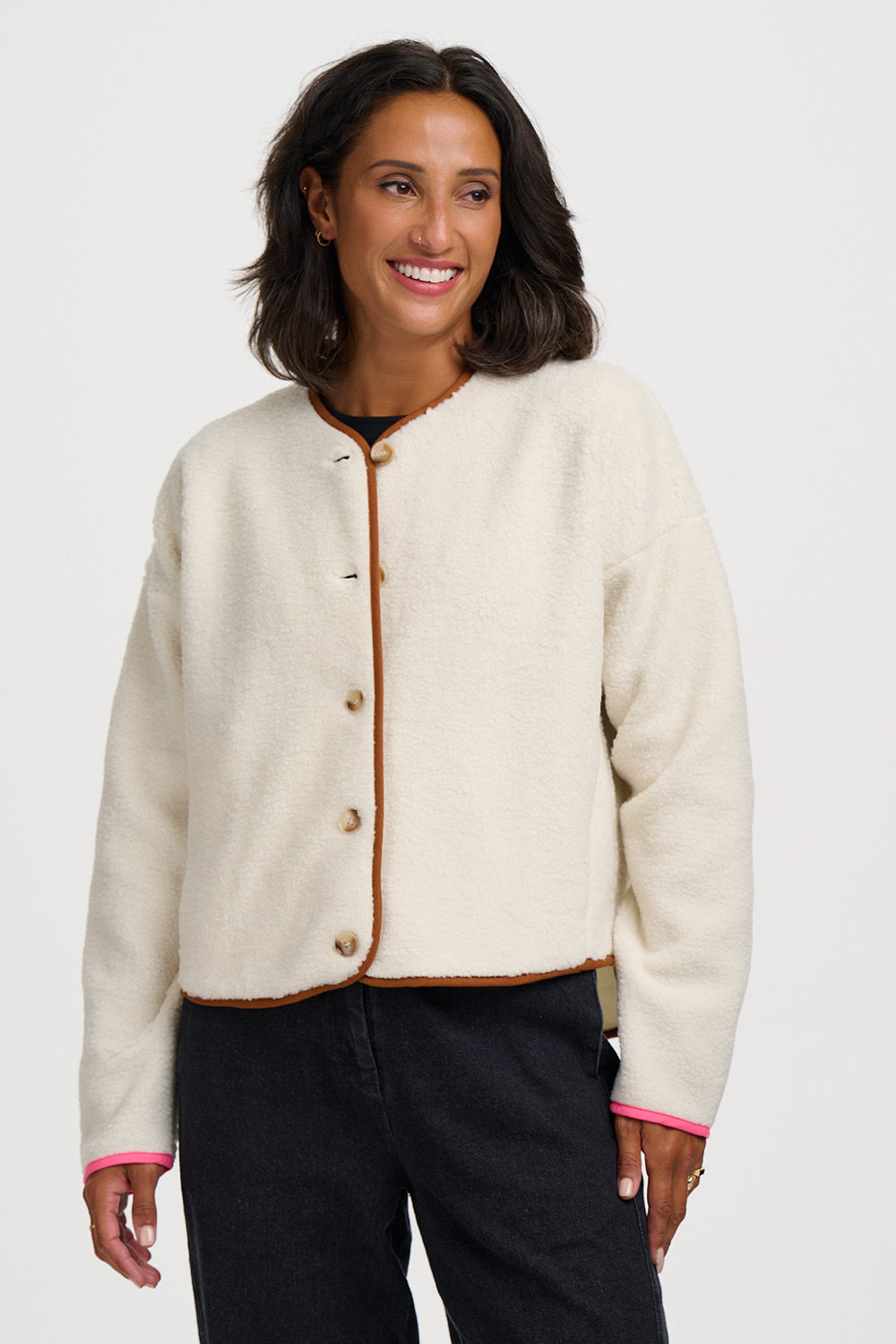 A woman wearing a cream-colored fuzzy cropped cardigan with brown trim and buttons, paired with black pants, against a light gray studio background.