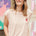 A woman smiling and wearing a pink sleeveless T-shirt with a rainbow parrot embroidery on the chest pocket, paired with blue jeans.