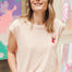 A woman smiling and wearing a pink sleeveless T-shirt with a rainbow parrot embroidery on the chest pocket, paired with blue jeans.