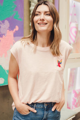 A woman smiling and wearing a pink sleeveless T-shirt with a rainbow parrot embroidery on the chest pocket, paired with blue jeans.