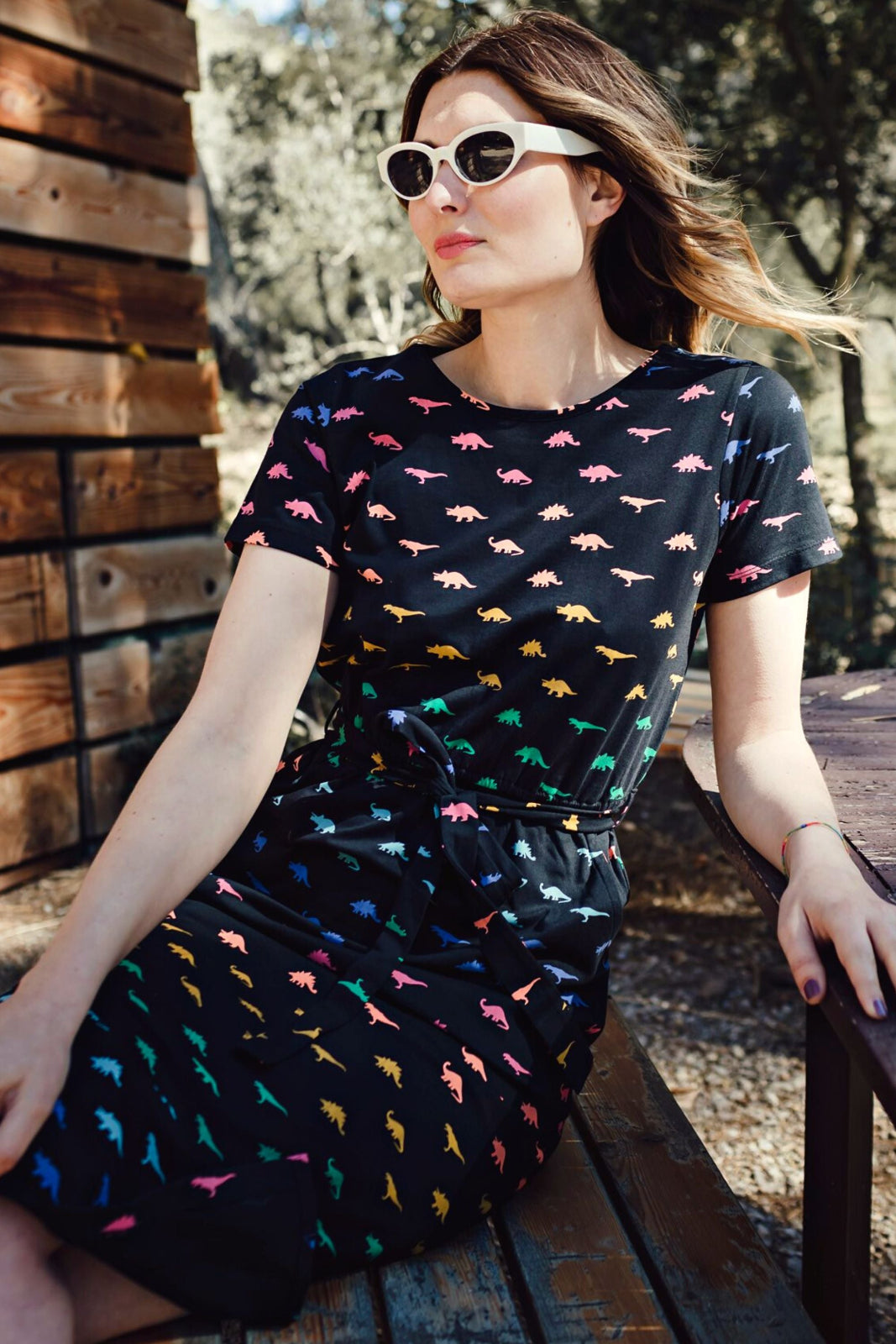 A person wearing a black midi length jersey dress with a colorful dinosaur print, paired with a matching tie belt and sunglasses.