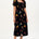 A model wearing a midi dress with a square neckline, short sleeves, and a shirred bust. The dress has a black background with a vibrant Tropical Parrots print in colorful patterns.