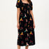A model wearing a midi dress with a square neckline, short sleeves, and a shirred bust. The dress has a black background with a vibrant Tropical Parrots print in colorful patterns.