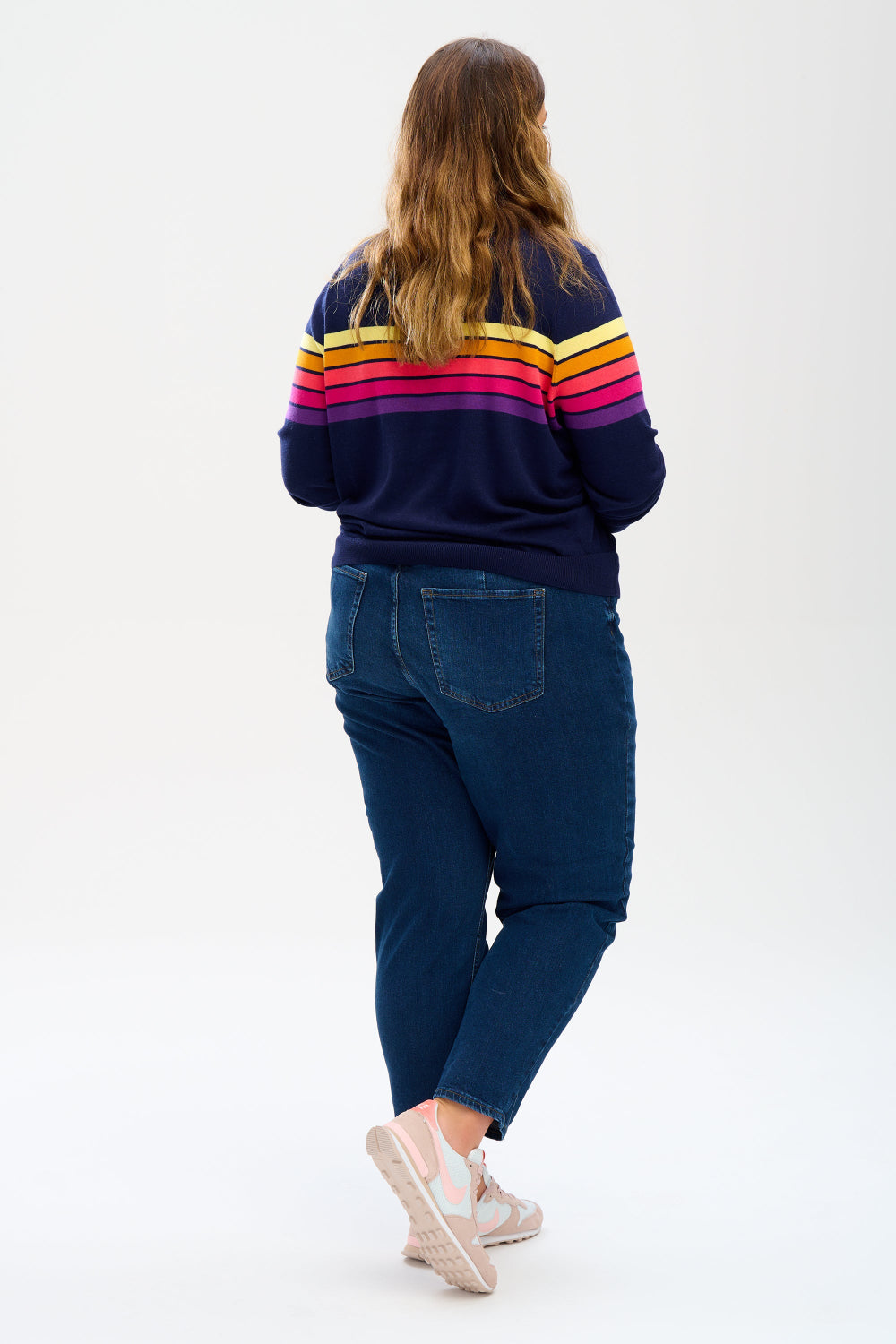 Sugarhill Brighton model wearing a navy blue jumper with horizontal retro stripe bands in pink, yellow, and other colours across the chest and shoulders.