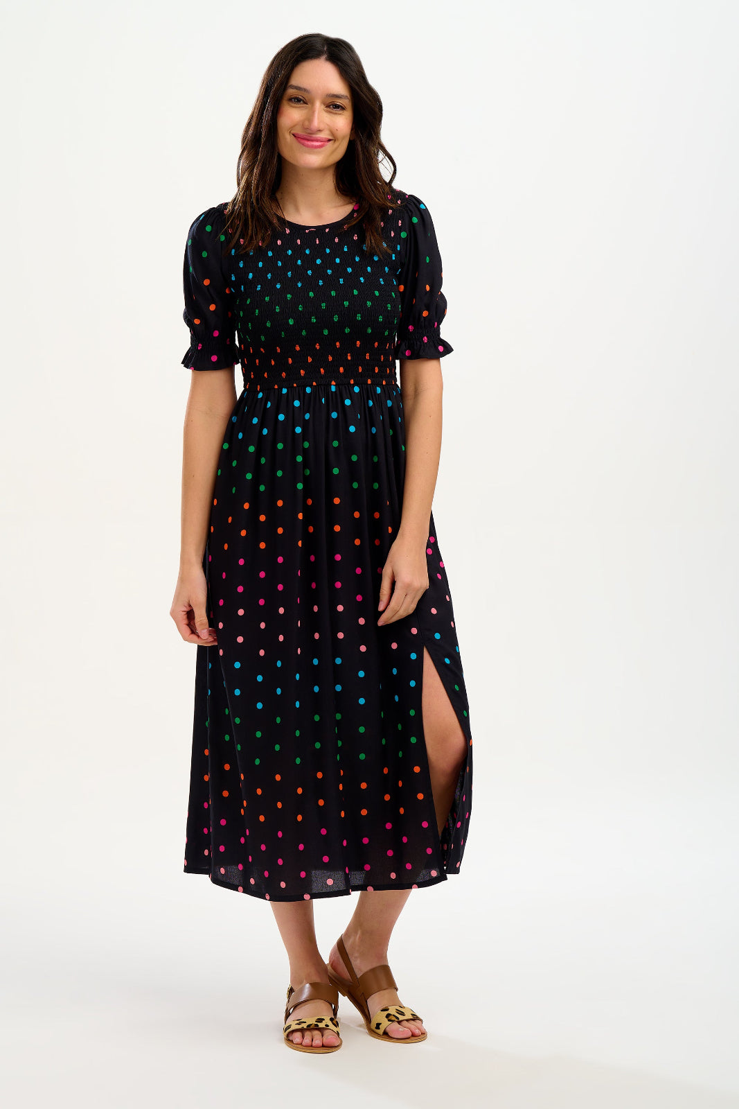 A model wearing a midi shirred dress with a black and rainbow polka print. The dress features a shirred bodice, elasticated puff sleeves, and a split hem.