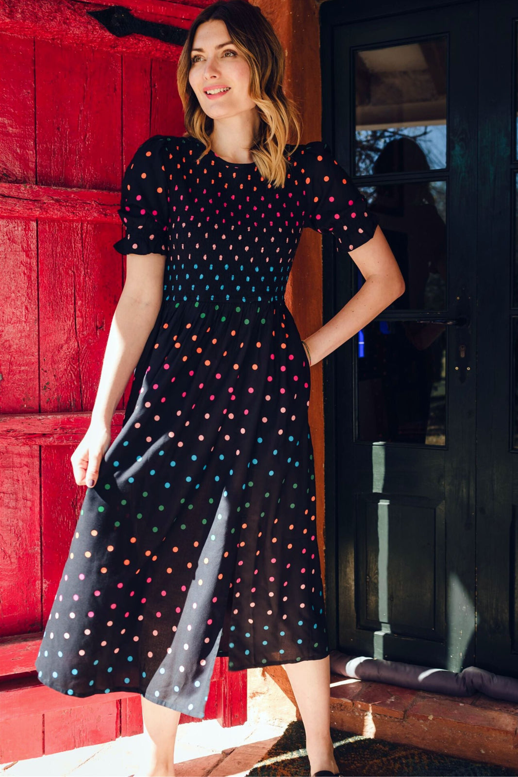 A model wearing a midi shirred dress with a black and rainbow polka print. The dress features a shirred bodice, elasticated puff sleeves, and a split hem.